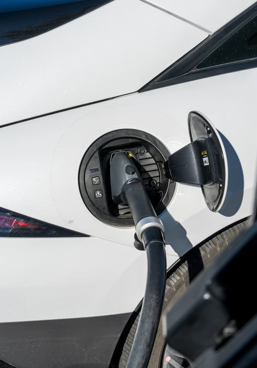 Photographie réaliste en extérieur d'une borne de recharge pour véhicule électrique montée sur une façade ou au sol, avec un technicien électricien en tenue professionnelle effectuant l'installation ou en train d'intervenir sur la borne avec ses outils. La borne doit être clairement visible et être le sujet principal de l'image. Le technicien peut être montré en train de vérifier les connexions, d'installer ou de finaliser la mise en service de la borne. Qualité professionnelle, cadrage mettant en évidence l'équipement et le professionnalisme du travail réalisé.